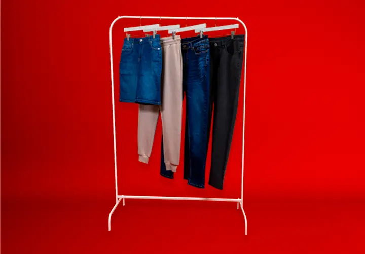 Wieszak prezentuje cztery pary spodni: jasnoniebieskie i czarne dżinsy, beżowe joggery oraz dżinsową spódnicę na tle intensywnie czerwonym.